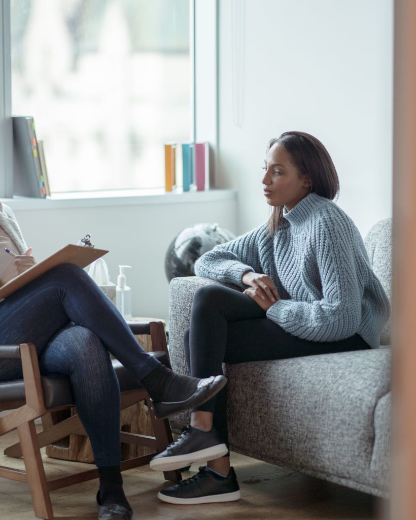 A Therapist meets with her female client in her office.  The client is seated on a sofa with her arms across her body as she looks visibly nervous.  The Therapist is seated in a chair in front of her as she talks about what to expect from the appointment and takes notes on her clipboard.